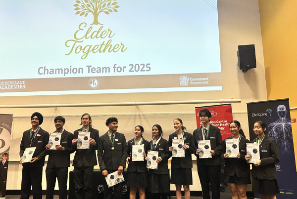 Group of high school students from Queensland Academies Health Sciences Campus standing on stage in uniform, holding certificates and medals, under a presentation screen that reads “Congratulations – Elder Together – Champion Team for 2025.