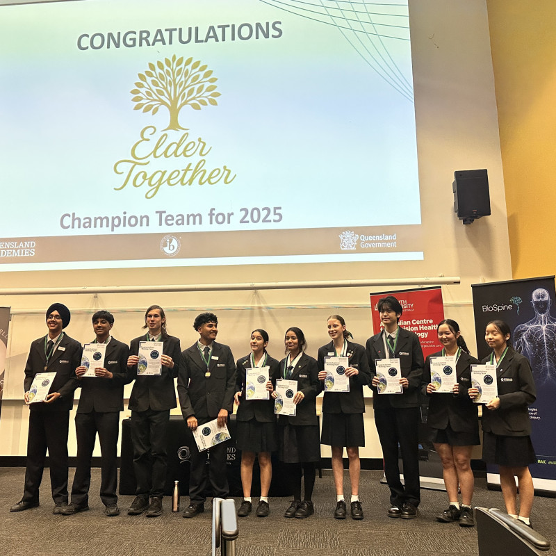 Group of high school students from Queensland Academies Health Sciences Campus standing on stage in uniform, holding certificates and medals, under a presentation screen that reads “Congratulations – Elder Together – Champion Team for 2025.