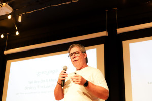 Person in a white ezylegal t-shirt and glasses speaking into a microphone in front of a screen showing the ezylegal platform, presenting to an audience.