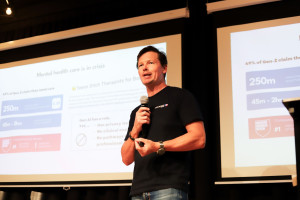 Man in a black t-shirt with Ascenda branding presenting on mental health care innovation, standing in front of a screen with slides showing statistics and information about Gen Z mental health needs.
