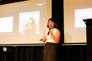 Woman in white sleeveless top and dark trousers presenting on stage with a microphone, standing in front of a screen displaying SmartAIConnect branding and a photo of her in the outdoors.
