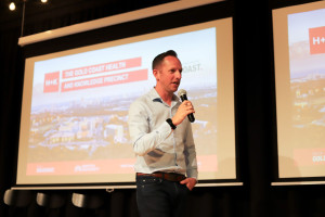 Aaron Isted, Senior Manager for Industry Growth, speaking into a microphone in front of a presentation slide displaying the Gold Coast Health and Knowledge Precinct branding.