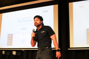 Man in a black Sophiie-branded polo shirt passionately presenting on stage with a microphone, standing in front of a large screen displaying the Sophiie AI virtual receptionist platform.