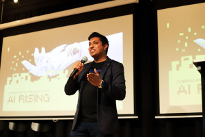 Man in a Zeligate t-shirt and blazer enthusiastically presenting with a microphone in hand, standing in front of an AI Rising event screen with robotic-themed graphics.