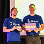 Two Cut Compass team members smile on stage while holding their People’s Choice Award certificates and mugs.