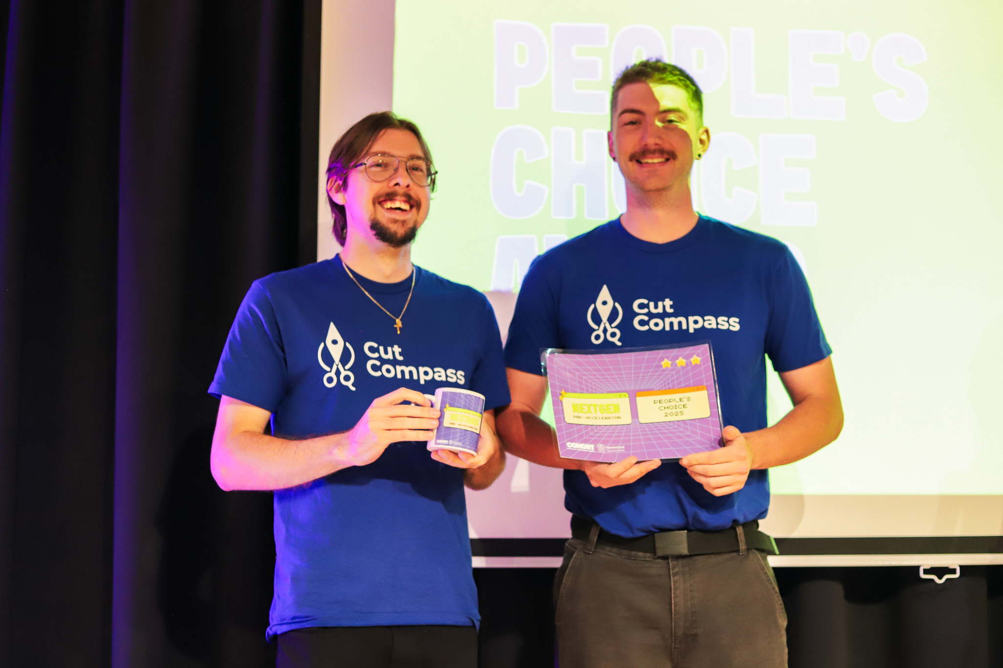 Two Cut Compass team members smile on stage while holding their People’s Choice Award certificates and mugs.