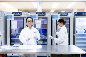 A researcher wearing a white Griffith University lab coat stands at a laboratory bench preparing a syringe, while another researcher retrieves supplies from a medical fridge behind her.