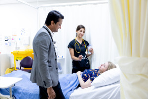 A doctor and nurse speak with a female patient lying in a hospital bed, with the nurse holding medical equipment and the doctor standing beside the bed.